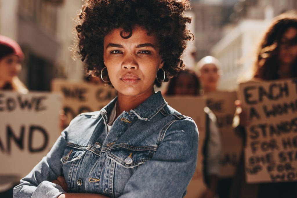 Woman stands in foreground, looking directly at camera, behind group of protesters.
